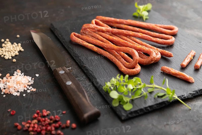 Traditional polish smoked pork sausage kabanos on a slate cutting board with salt and pepper on black concrete background. Side view, close up, selective focus
