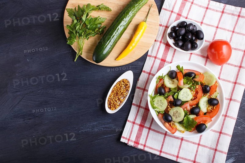 Vegetarian salad of tomatoes, cucumbers, parsley, olives and mustard on linen tablecloth and black wooden background, top view, flat lay
