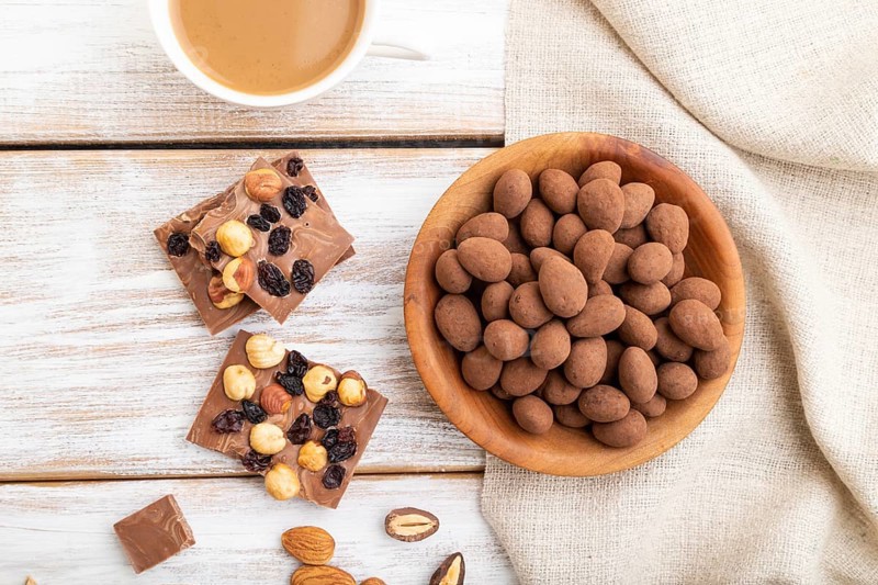 Almond in chocolate dragees in wooden plate and a cup of coffee on white wooden background and linen textile. Top view, close up, flat lay