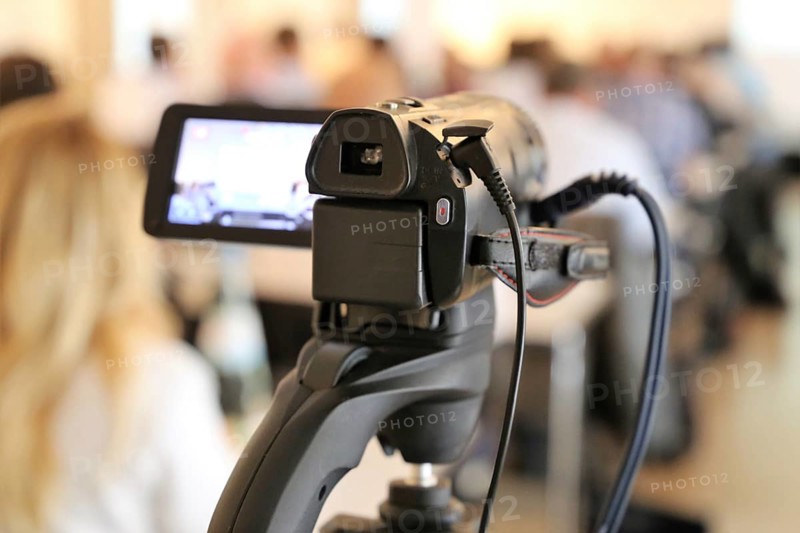 Close-up of a camcorder at a conference or press conference. The guests can be recognised out of focus in the background
