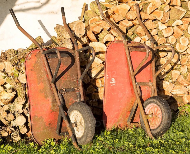 Two old red metal wheelbarrows leaning against pile wood logs in garden, Cherhill, Wiltshire, England, UK