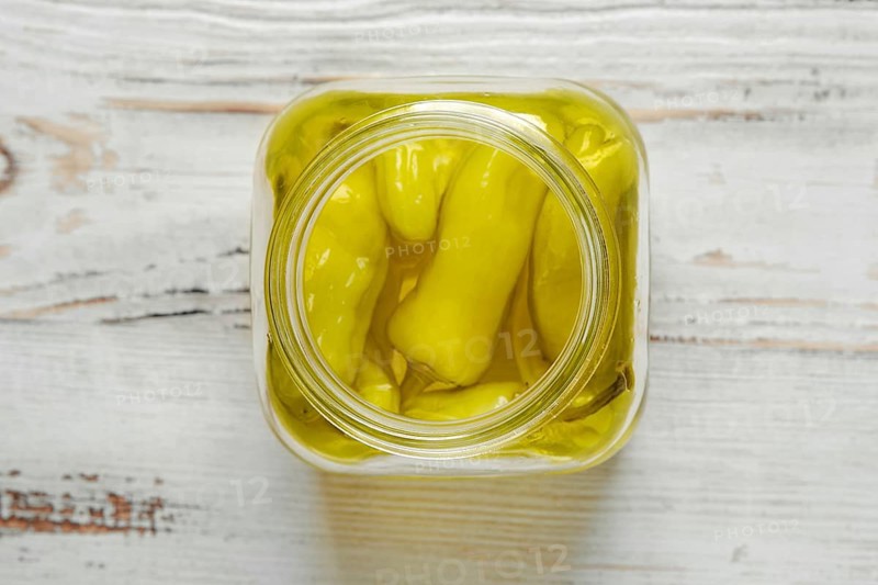 A glass jar of pickled peppers sits on a white wooden table. The jar is filled with whole, yellow peppers that have been preserved in oil or brine