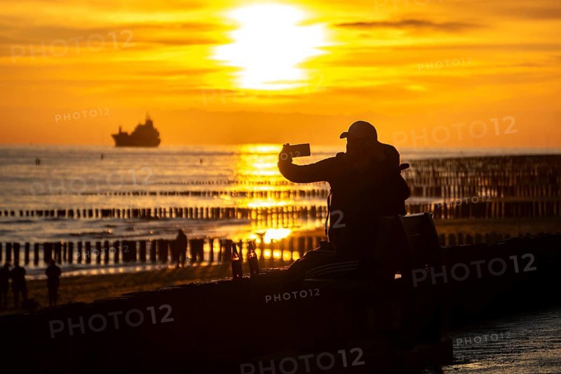 Sunset on the beach of Zoutelande, beach with wooden pile breakwaters, tourists, cargo ship sailing towards the Westerschelde, Zeeland, Netherlands
