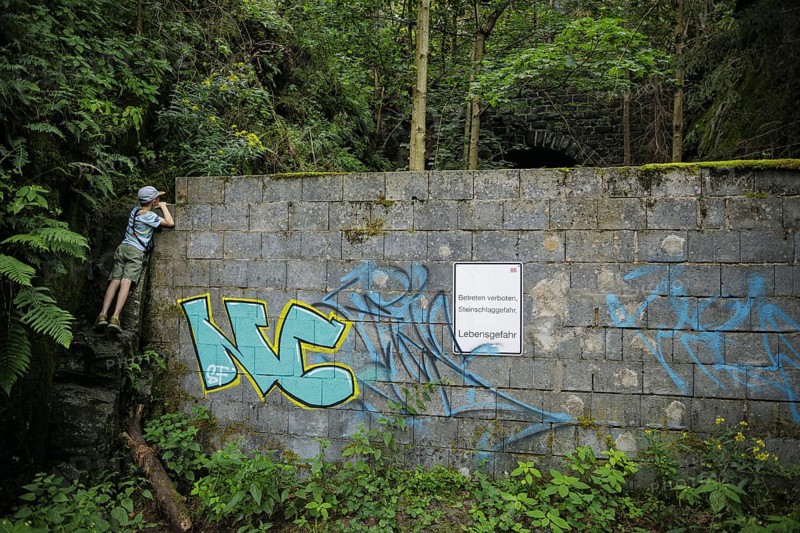 Symbolic photo on the subject of curiosity in children. A boy stands on a stone and looks over a wall with a sign saying 'Äô No trespassing, danger to life'Äô. Lichtenberg, 21.07.2024