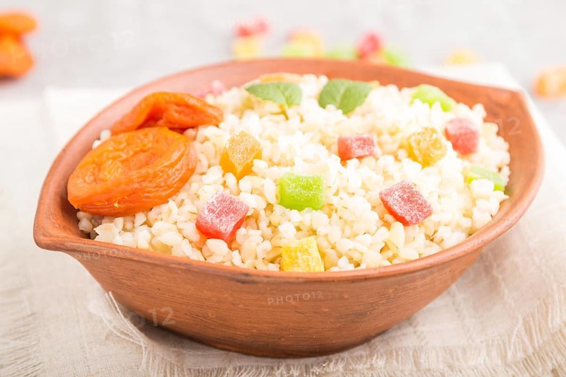 Bulgur porridge with dried apricots and candied fruits in clay bowl on a gray concrete background and linen textile. Side view, close up, selective focus. Turkish traditional cuisine