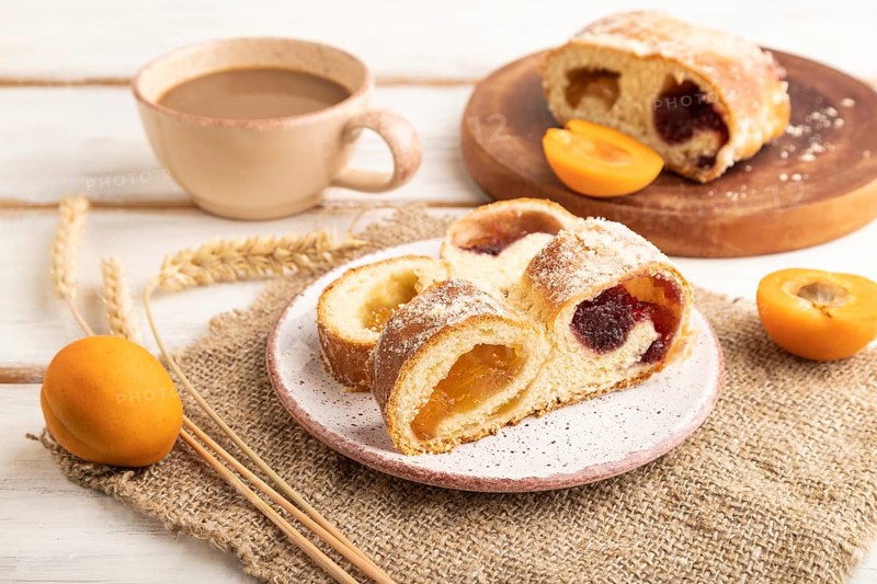 Homemade sweet bun with apricot jam and cup of coffee on white wooden background and linen textile. side view, close up