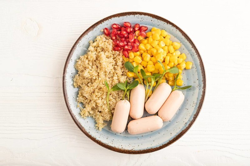 Mixed quinoa porridge, sweet corn, pomegranate seeds and small sausages on white wooden background. Top view, flat lay, close up. Food for children, healthy food concept