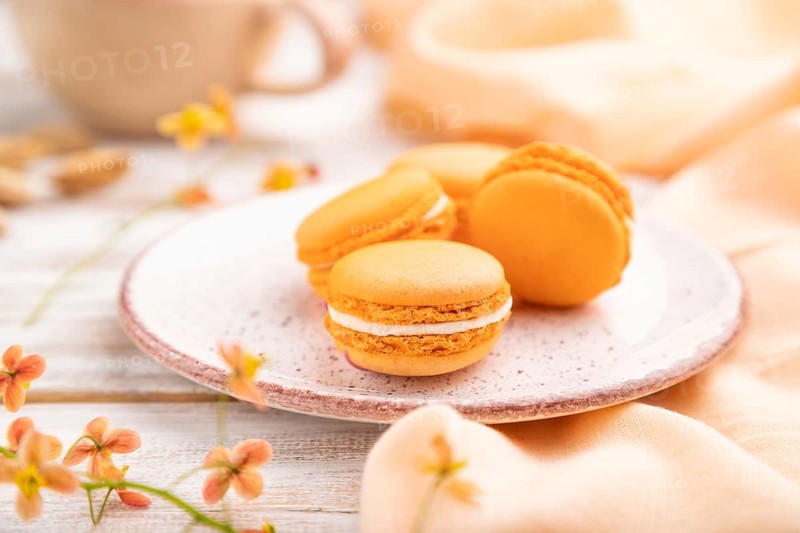 Orange macarons or macaroons cakes with cup of apricot juice on a white wooden background and orange linen textile. Side view, close up, selective focus