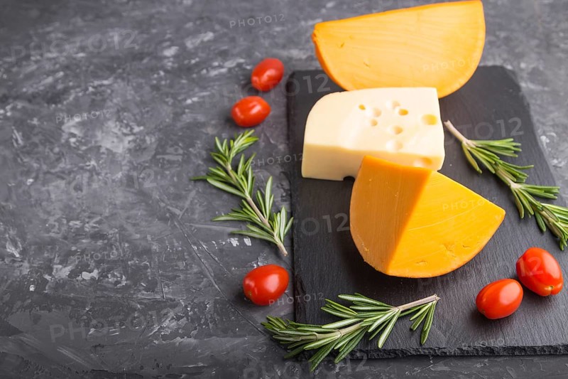 Various types of cheese with rosemary and tomatoes on black slate board on a black concrete background. Side view, close up, copy space, selective focus