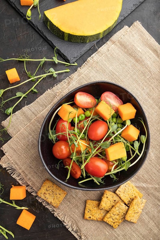 Vegetarian vegetable salad of tomatoes, pumpkin, microgreen pea sprouts on black concrete background and linen textile. Top view, flat lay, close up