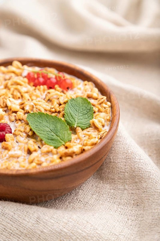 Wheat flakes porridge with milk, raspberry and currant in wooden bowl on white wooden background and linen textile. Side view, close up, selective focus