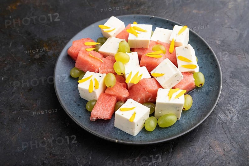 Vegetarian salad with watermelon, feta cheese, and grapes on blue ceramic plate on black concrete background. Side view, close up