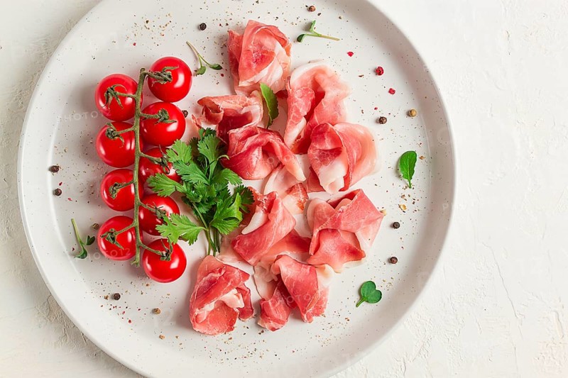 Sliced prosciutto, appetizer, with cherry tomatoes, on a white plate, top view
