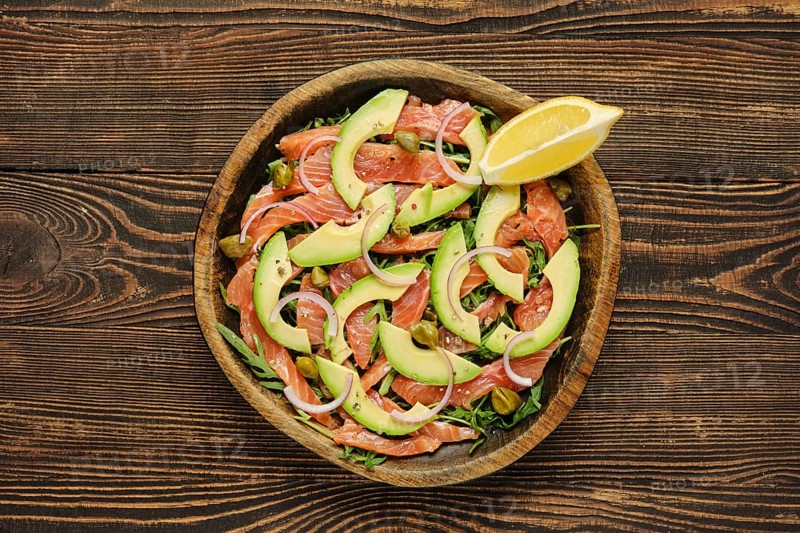 Salad with slices of fresh salmon, creamy avocado, and crisp onions, artfully arranged in a wooden bowl. It is complemented by a wedge of lemon, adding a refreshing touch
