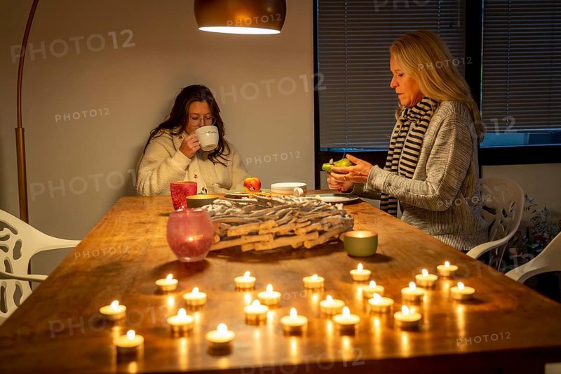 Symbolic image energy saving, cold winter, energy crisis, cold flat, mother and daughter dressed warmly at dinner, only one lamp on, candlelight, heating on to a minimum