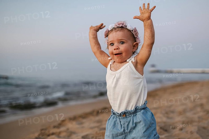 A baby in summery clothes and flowers in her hair stands on the beach at sunset and stretches both arms upwards