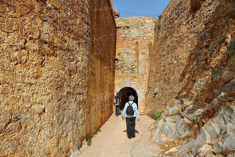 Two people walking through a narrow passageway between high stone walls of an ancient fortress, Venetian Sea Fortress, Leprosy Island, Spinalonga, Elounda Bay, Mirabello Bay, East Crete, Crete, Greek Islands, Greece, Europe