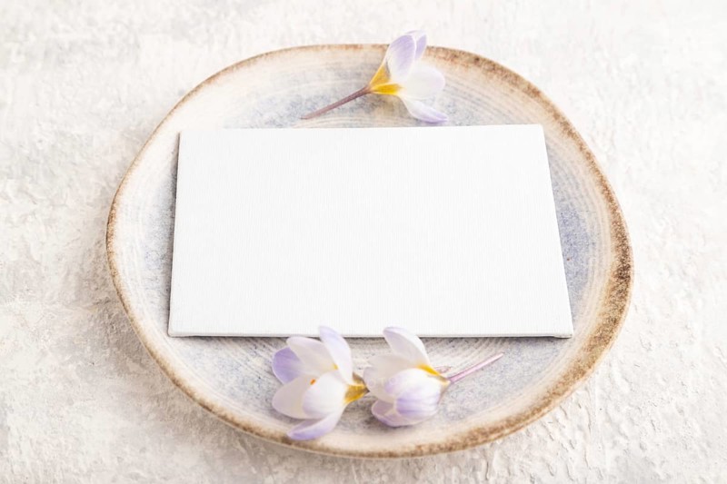 White paper invitation card, mockup with crocus flowers on ceramic plate and gray concrete background. Blank, flat lay, top view, still life, copy space, wedding invitation