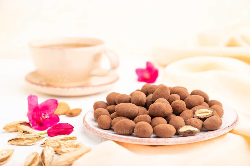 Almond in chocolate dragees on ceramic plate and a cup of coffee on white concrete background and orange linen textile. Side view, close up, selective focus