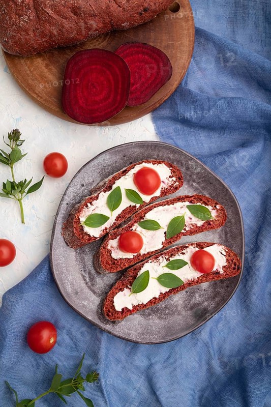 Red beet bread sandwiches with cream cheese and tomatoes on white concrete background and blue linen textile. top view, flat lay, close up