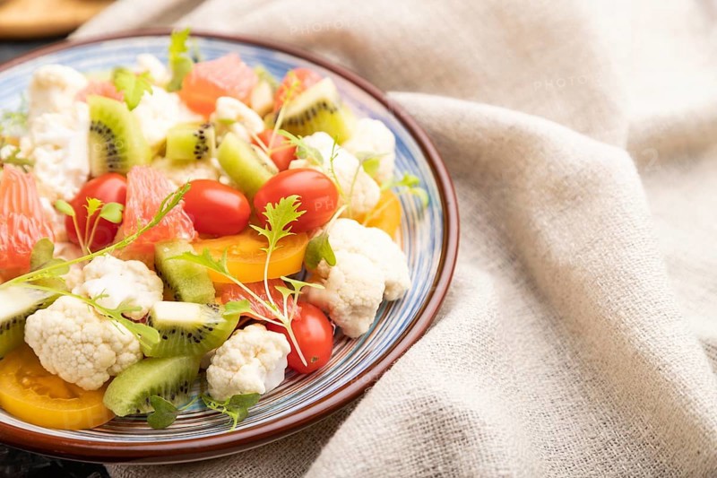 Vegetarian salad of cauliflower cabbage, kiwi, tomatoes, microgreen sprouts on black concrete background and linen textile. Side view, close up, selective focus