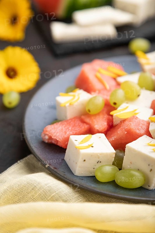 Vegetarian salad with watermelon, feta cheese, and grapes on blue ceramic plate on black concrete background and yellow linen textile. Side view, close up, selective focus