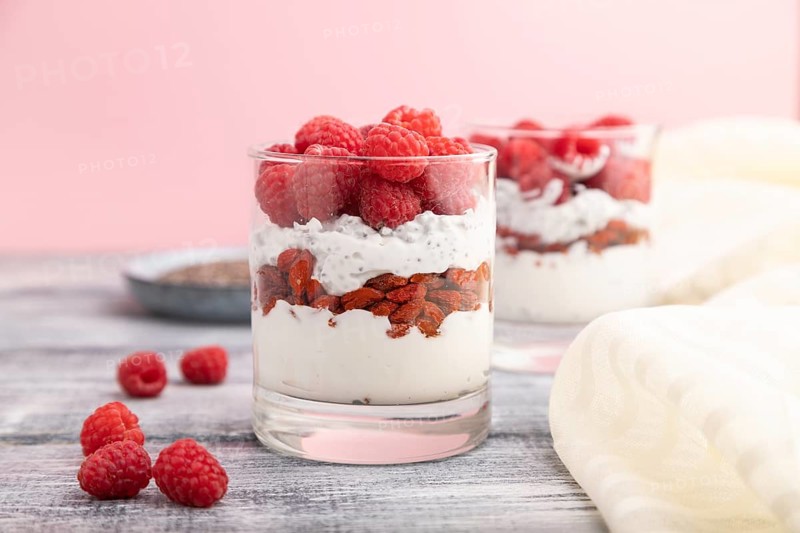 Yogurt with raspberry, goji berries and chia seeds in glass on gray wooden background and linen textile. Side view, close up, selective focus