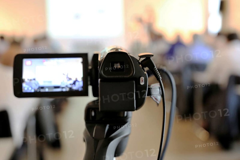 Close-up of a camcorder at a conference or press conference. The guests can be recognised out of focus in the background