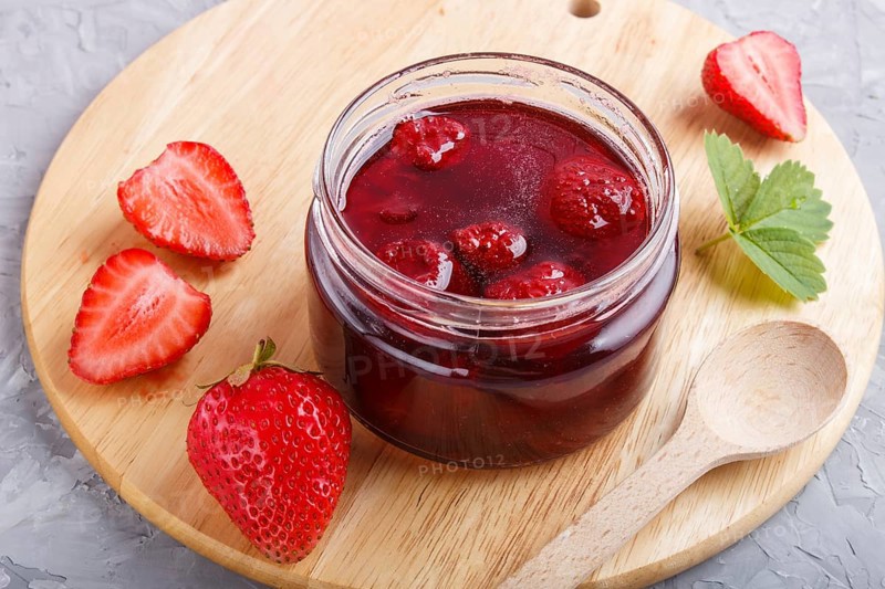 Strawberry jam in a glass jar with berries and leaves on gray concrete background. Homemade, close up, side view, selective focus