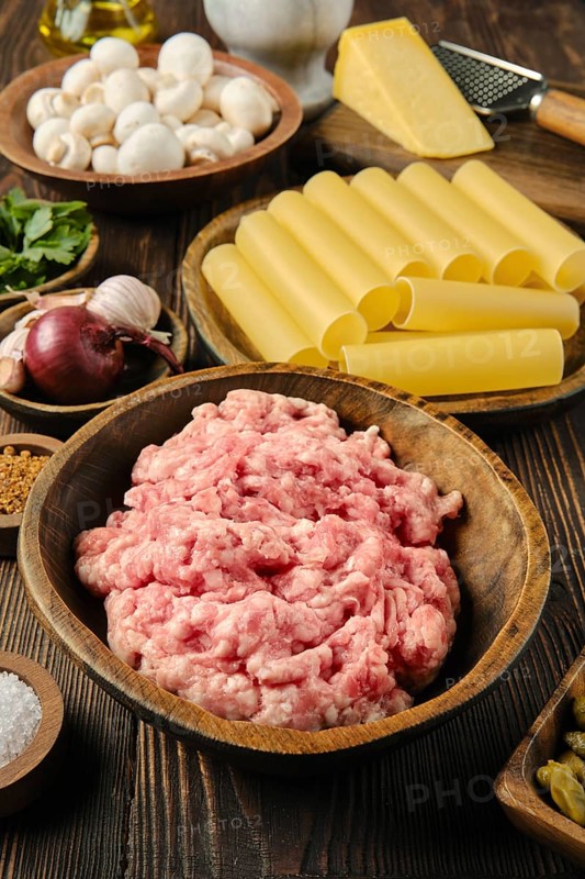 A wooden bowl filled with raw ground meat is surrounded by pasta, mushrooms, garlic, and cheese on a rustic wooden table, showcasing a vibrant array of fresh cooking ingredients