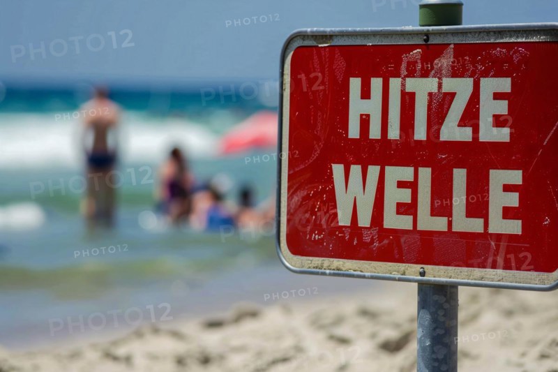 Warning sign with German text 'Hitzewelle' (Heat wave) in front of blurry sunny beach with people. KI generiert, generiert, AI generated