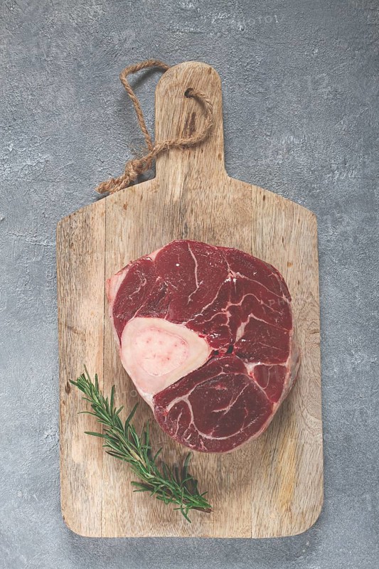 A shank on the bone, raw meat for ossobuco, on a wooden chopping board, with a branch of rosemary, top view, no people