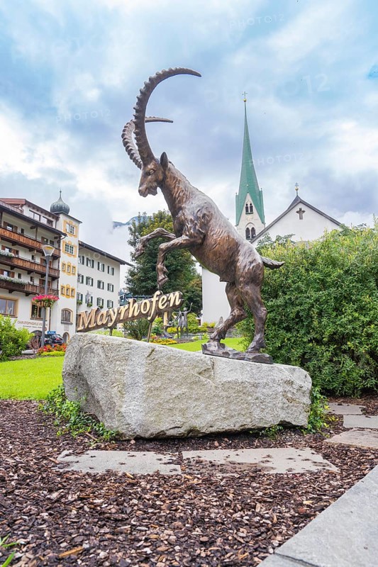 Bronze statue of a rising goat in front of a church and buildings in the background, Mayrhofen, Zillertal, Austria, Europe
