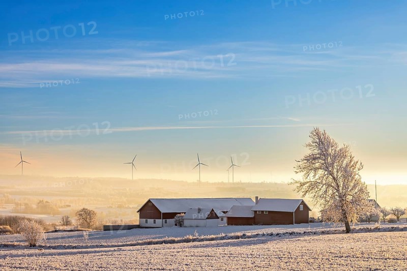Farmhouse in a field with snow and frost and low cold fog in a valley and wind turbines on the horizon in the wintry countryside, Sweden, Europe