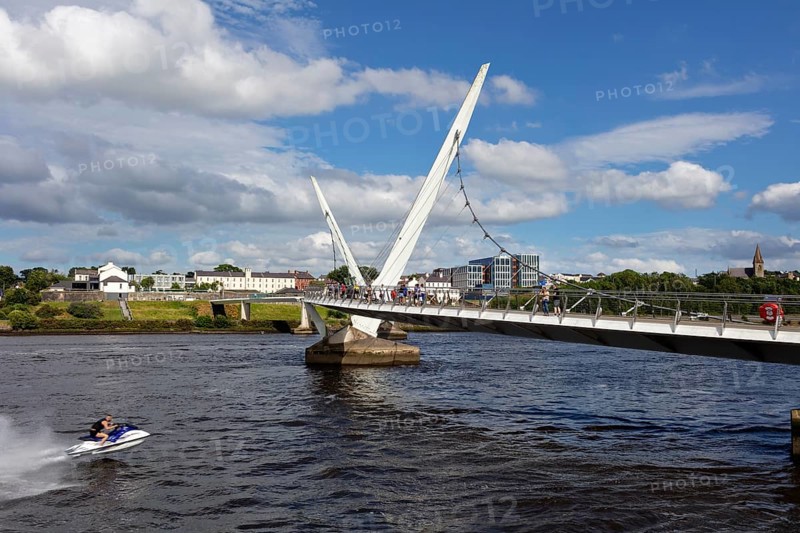 A jet ski passes under a striking bridge surrounded by calm waters, Londonderry