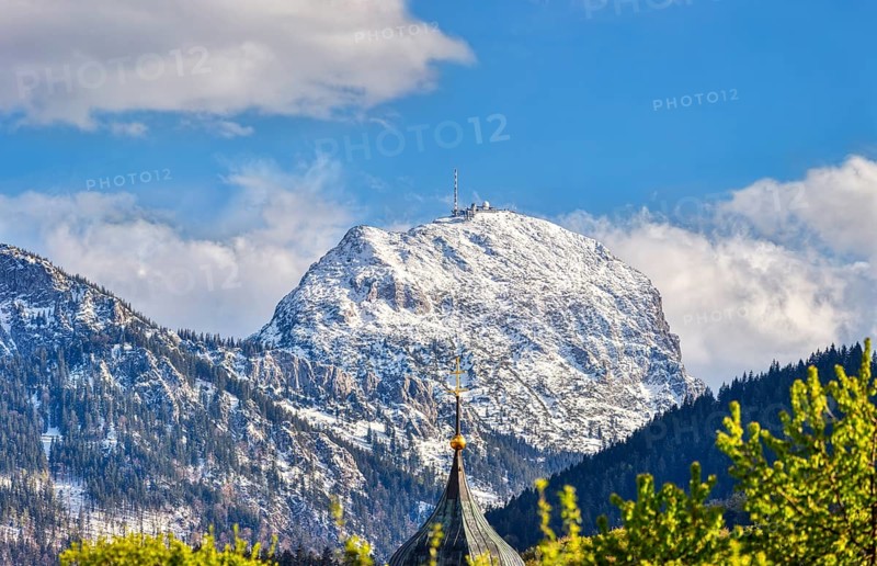 Snow-covered mountain peak under a blue sky with clouds, in the foreground a church tower and green tree tops, Wendelstein