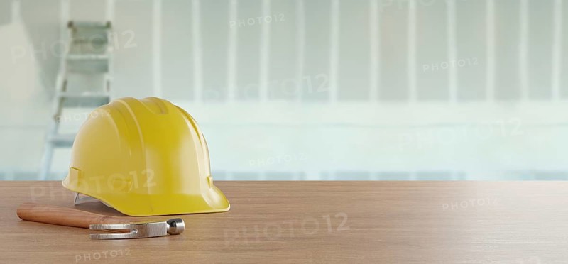 Yellow hard hat and hammer resting on a wood surface in front of drywall and ladder banner