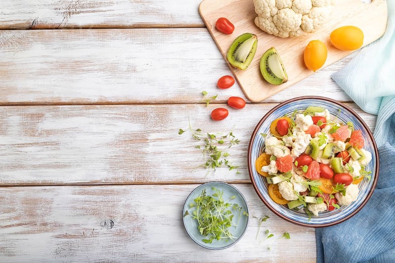 Vegetarian salad of cauliflower cabbage, kiwi, tomatoes, microgreen sprouts on white wooden background and blue linen textile. Top view, flat lay, copy space
