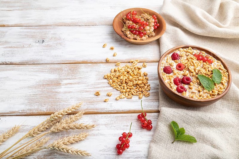 Wheat flakes porridge with milk, raspberry and currant in wooden bowl on white wooden background and linen textile. Side view, copy space