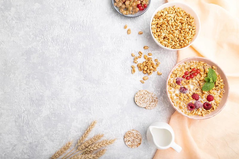 Wheat flakes porridge with milk, raspberry and currant in ceramic bowl on gray concrete background and orange linen textile. Top view, flat lay, copy space