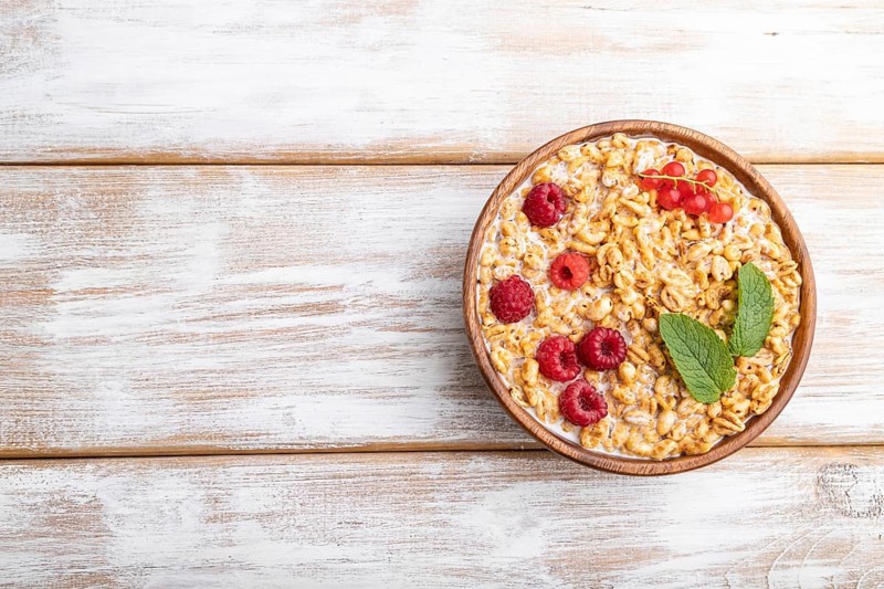 Wheat flakes porridge with milk, raspberry and currant in wooden bowl on white wooden background. Top view, flat lay, copy space