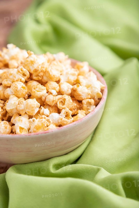 Popcorn with caramel in ceramic bowl on brown concrete background and green textile. Side view, close up, selective focus