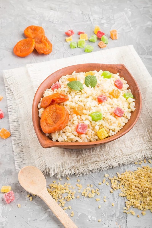 Bulgur porridge with dried apricots and candied fruits in clay bowl on a gray concrete background and linen textile. Side view, close up. Turkish traditional cuisine