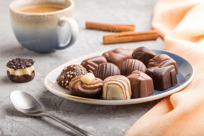 Different chocolate candies and a cup of coffee on a gray concrete background and orange textile. side view, close up, selective focus