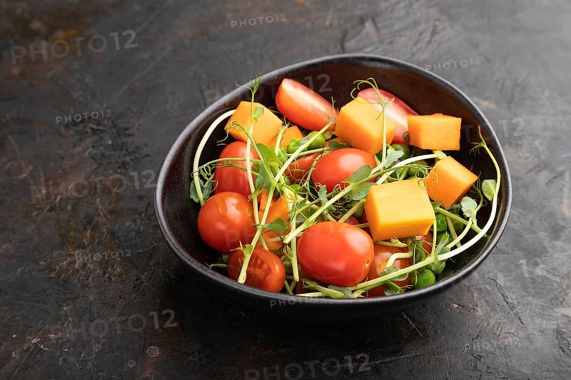 Vegetarian vegetable salad of tomatoes, pumpkin, microgreen pea sprouts on black concrete background. Side view, copy space, close up