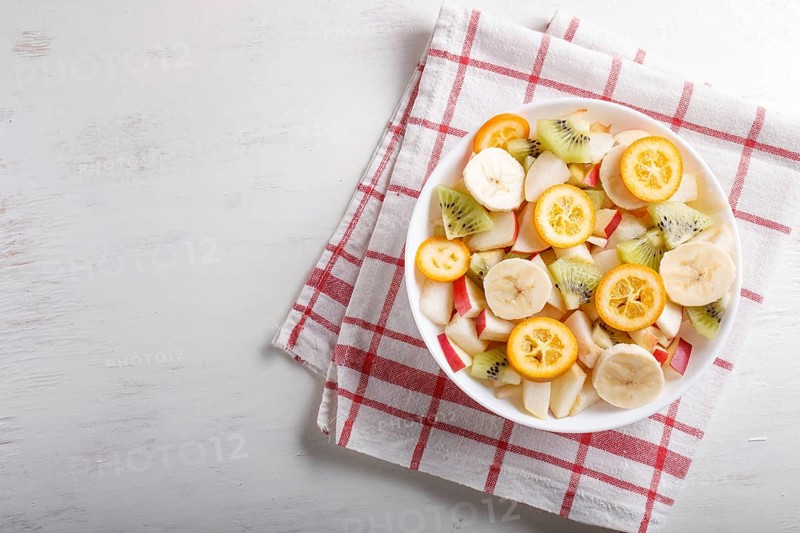 Vegetarian salad of bananas, apples, pears, kumquats and kiwi on linen tablecloth, top view, flat lay, copy space. white wooden background
