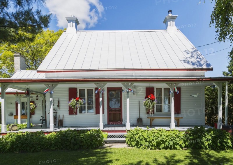 Old circa 1840 white horizontal wood plank with red trim Canadiana style house facade with silver standing seam sheet metal roof in early summer, Quebec, Canada, North America