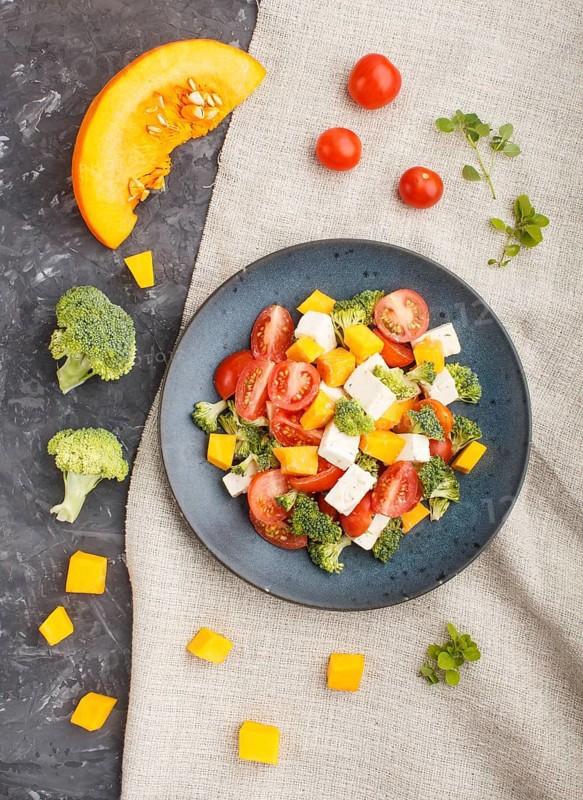 Vegetarian salad with broccoli, tomatoes, feta cheese, and pumpkin on a blue ceramic plate on a black concrete background and linen textile, top view, close up, flat lay