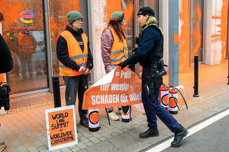 Police officer confiscates Last Generation banner after colour attack on Total Energies building. The banner reads: LNG = profit for a few, harm for all!, on a sign: World LNG summit = worldwide catastrophe, Berlin, 6 November 2024