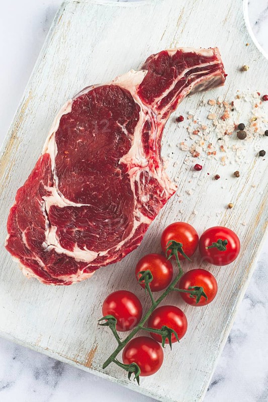 Raw rib eye steak, cowboy steak, on a white wooden chopping board, marble table, top view, no people